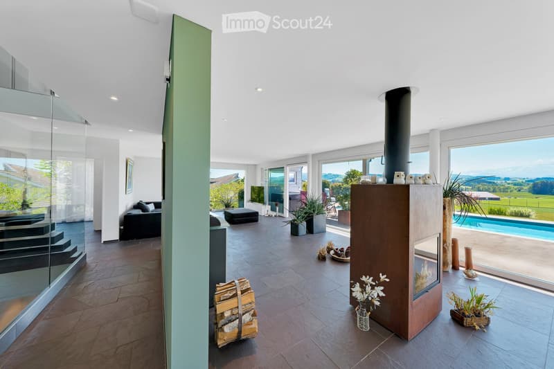 Modern living room with large windows offering scenic views, a sleek black sofa, an ottoman, a contemporary fireplace, decorative vases, potted plants, and a stack of firewood.