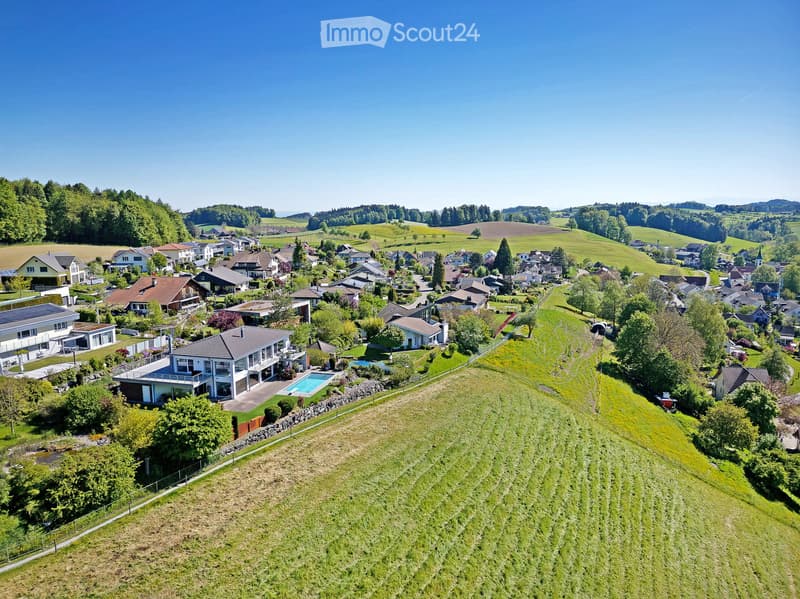 Aerial view of a suburban neighborhood with houses, gardens, and a swimming pool in the foreground, surrounded by green fields and trees.