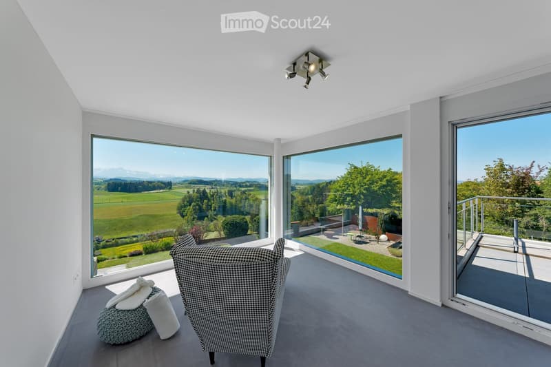 Bright living space with large windows offering scenic views, featuring a patterned armchair, a round stool with pillows, and modern ceiling lighting.