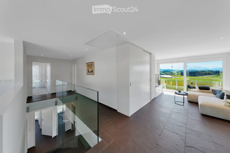 Modern living room with large windows offering scenic views, a glass staircase leading to another level, and contemporary furniture including a sofa, coffee table, and side table.