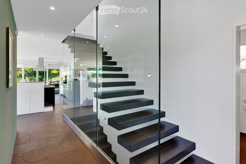 Modern hallway with a sleek glass staircase featuring dark wooden steps, white walls, and recessed lighting. The floor is made of polished wood tiles, and the space is open and airy with a view into the adjacent living area.