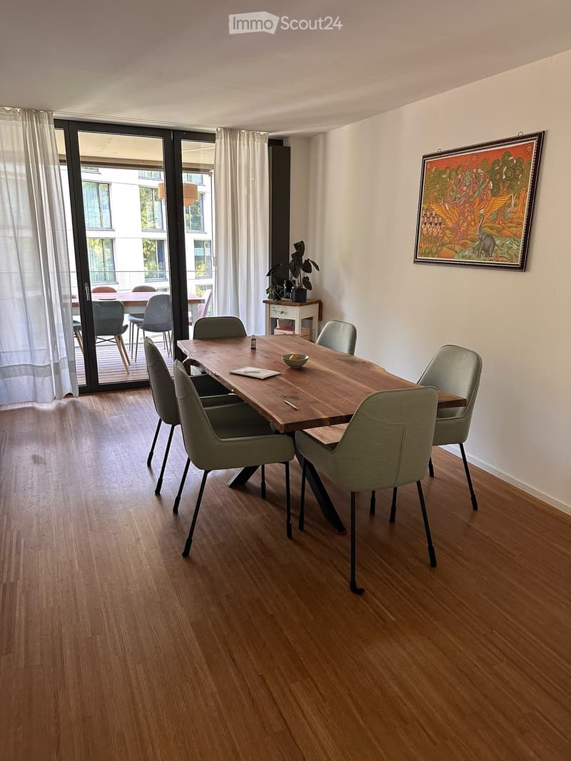 Dining room with wooden floor, large wooden dining table, six modern chairs, small plant on side table, and artwork on wall. Access to balcony through large sliding doors.