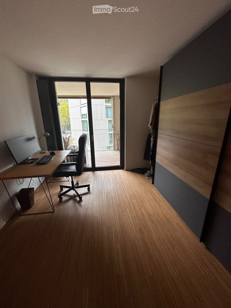 A modern, minimalist office space with wooden flooring, a large window, and a desk setup including a screen, keyboard, mouse, and a chair.