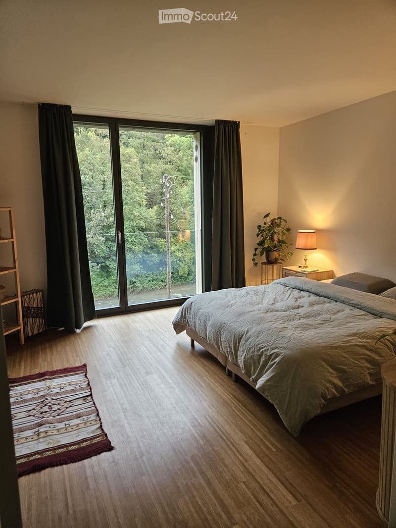 Modern bedroom with wooden flooring, large sliding glass door leading to outdoor view, bed with grey bedding, nightstand with lamp, small plant, and patterned rug.