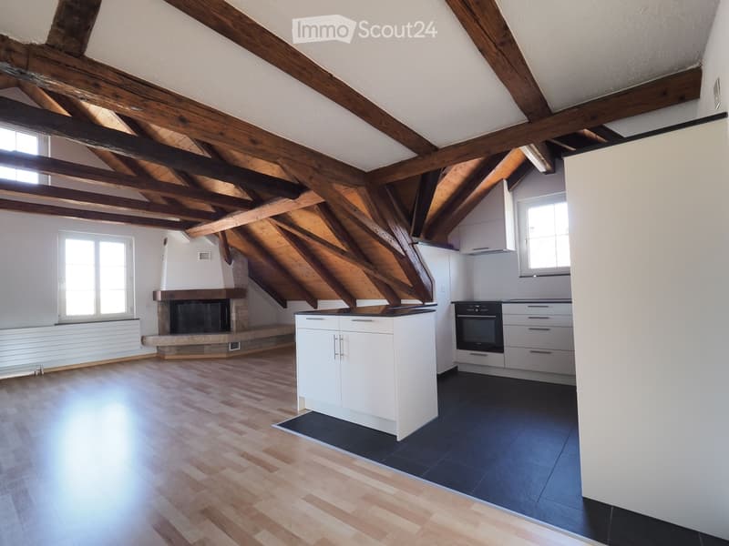 Open-plan attic space with exposed wooden beams, wooden flooring, a fireplace, a kitchen island, and built-in cabinets.