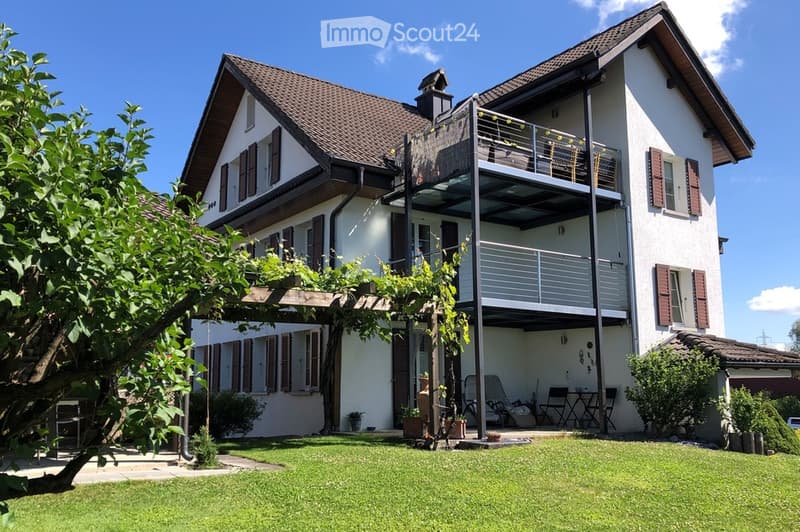 Two-story house with white exterior, brown roof, and balcony on the upper level. The house features multiple windows with shutters, a well-maintained lawn, and outdoor seating area.