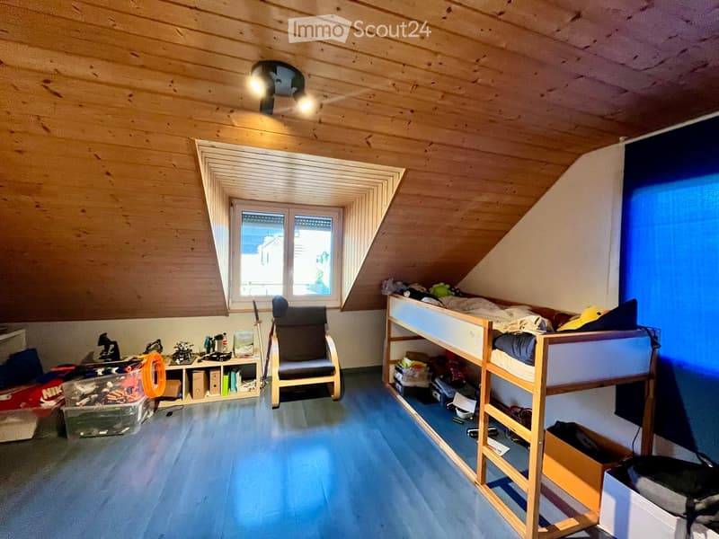 Cozy attic bedroom with wooden walls and ceiling, featuring a bunk bed, a wooden chair, a small table with various items, and storage boxes.