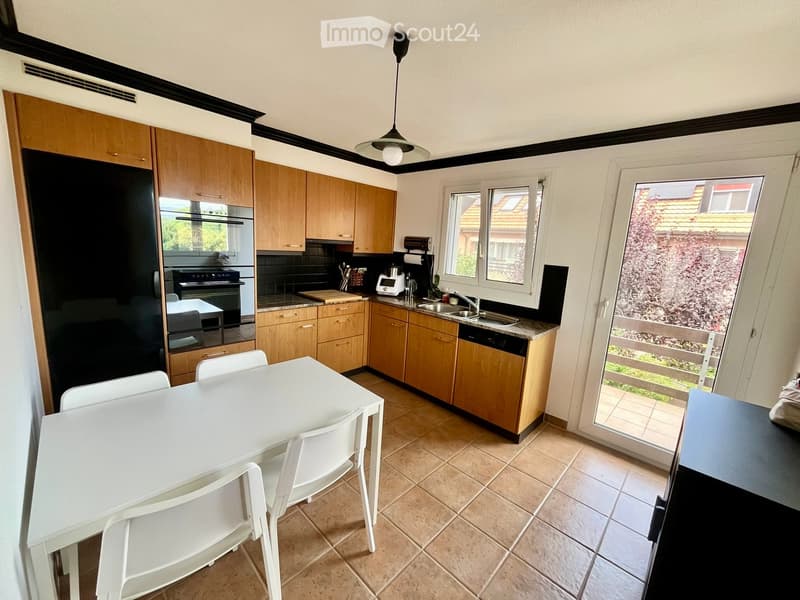 Modern kitchen with wooden cabinets, stainless steel appliances, a white dining table with chairs, and a large window overlooking a garden.