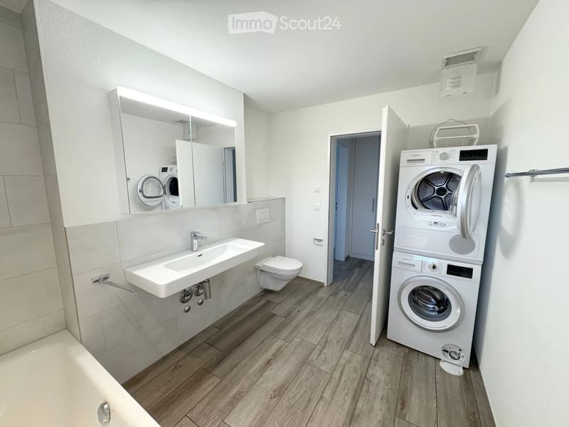 Modern bathroom with a sink, toilet, mirror, and a stacked washer and dryer unit. The space features light-colored tiles and wooden flooring.