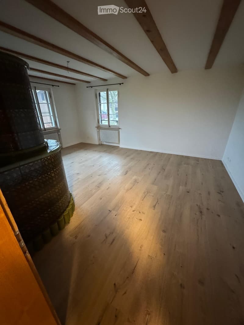 Empty room with wooden flooring, white walls, exposed wooden beams on the ceiling, and two windows with white frames. A radiator is visible beneath one window.