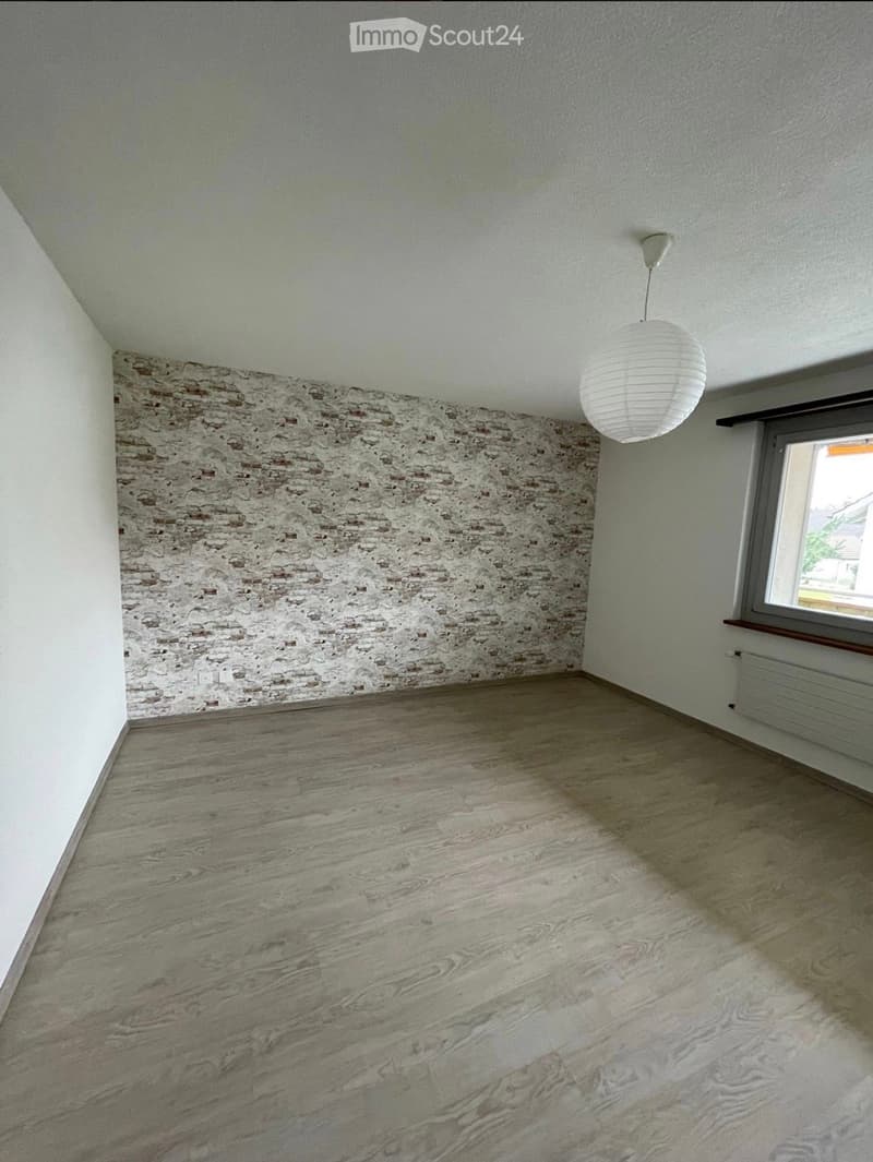 Empty room with wooden flooring, exposed brick wall, white pendant light, and a window with a view.