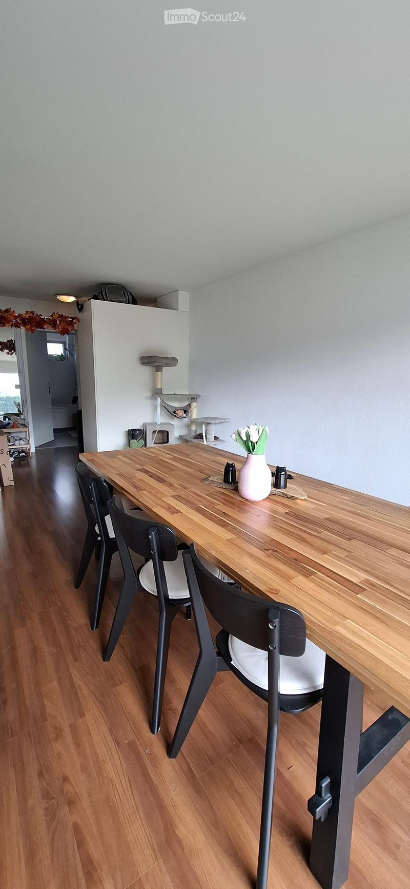A modern dining area with a wooden table, black chairs, a vase with white flowers, and a cat tree in the corner.