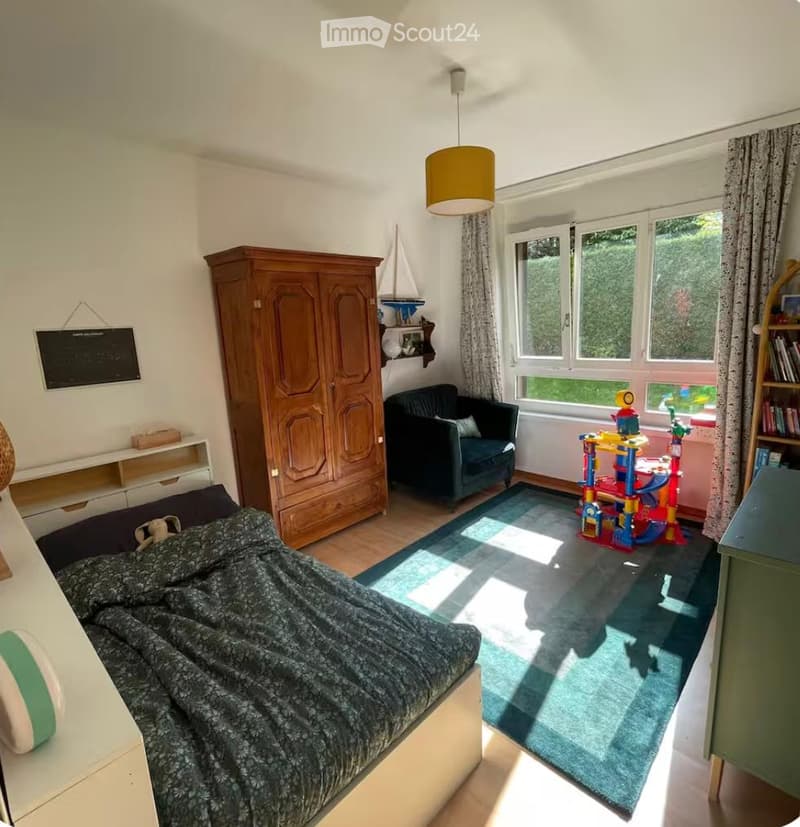Cozy bedroom with a double bed, dark green sofa, large wooden wardrobe, colorful toy structure, bookshelf, and a hanging lamp.