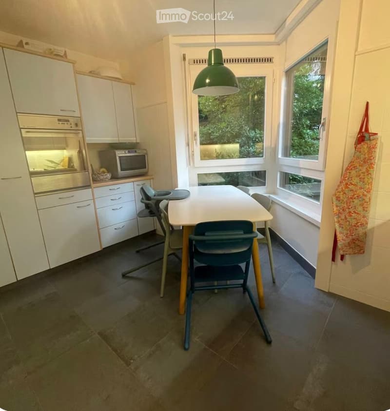 Bright kitchen with white cabinets, stainless steel oven, microwave, and a dining table with chairs, including a baby chair. Large windows provide natural light.