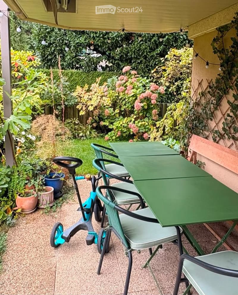 A small outdoor patio area with a green table and matching chairs, surrounded by lush greenery and potted plants. A small tricycle is visible near the edge of the patio.