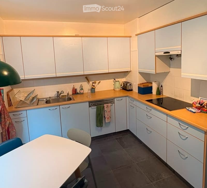 Bright kitchen with light blue cabinets, wooden countertops, and stainless steel appliances including a dishwasher and induction stove. Features a sink, faucet, and a small dining area with a table and chairs.