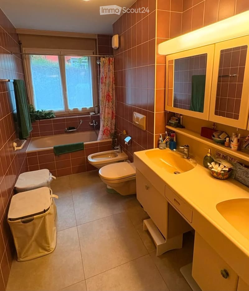 Bathroom with brown tiled walls and floor, featuring a bathtub, toilet, bidet, sink with mirror and cabinet, and a window with a shower curtain.