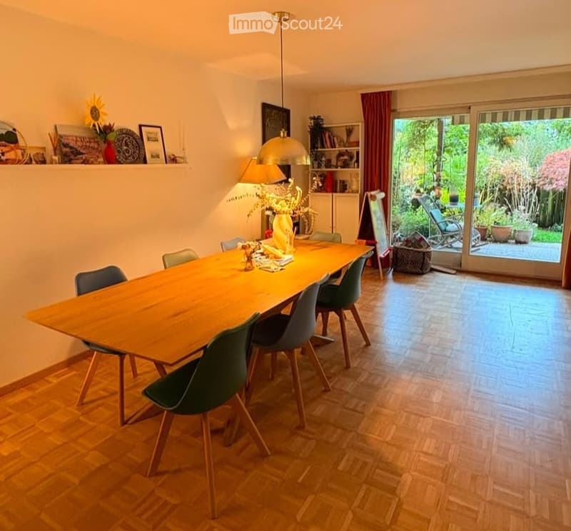Dining room with a long wooden table surrounded by green chairs, a hanging pendant light, and a shelf with decorative items.