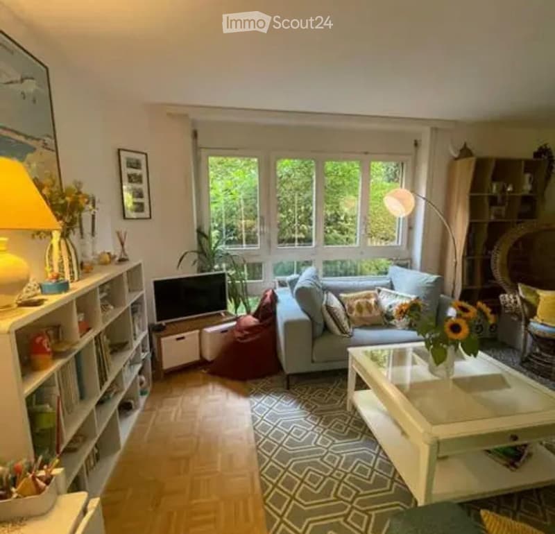Bright living room with large windows, light grey sofa, coffee table, bookshelf, floor lamp, and decorative items.