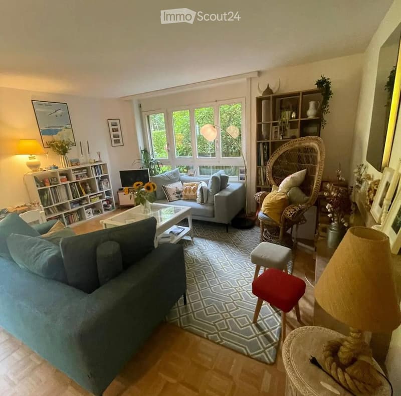Bright living room with large windows, two sofas, coffee table, bookshelf, wicker chair, and decorative items.