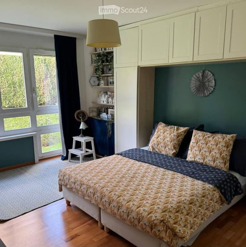 Cozy bedroom with a large bed, patterned bedspread, pillows, a small white stool, a lamp, built-in white cabinets, and a decorative wall piece. The room features wooden flooring and large windows with dark curtains.