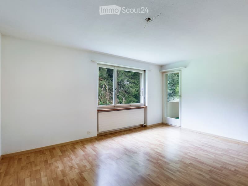 Empty room with light-colored wooden flooring, white walls, and large windows with a view of greenery. A radiator is visible below one window, and a door leads to another space.