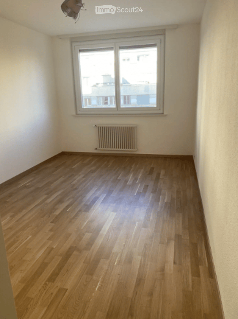 Completely empty room with wooden flooring, white walls, a single window, and a radiator below it.