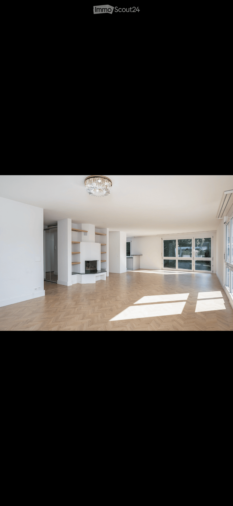 Empty room with wooden herringbone flooring, white walls, built-in fireplace with shelves, large windows, and a chandelier.