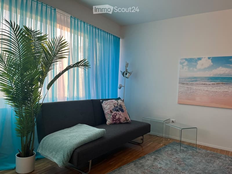 A modern living room with a grey sofa, decorative pillows, a small glass coffee table, a floor lamp, a large painting of a beach scene, and a potted plant near the window with blue curtains.