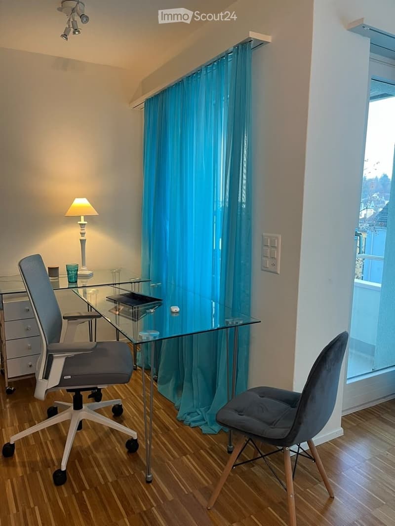 A modern, well-lit room with a glass desk, ergonomic chair, and a comfortable seating chair. The room features wooden flooring, a blue curtain, and a small table lamp, creating a cozy and functional workspace.