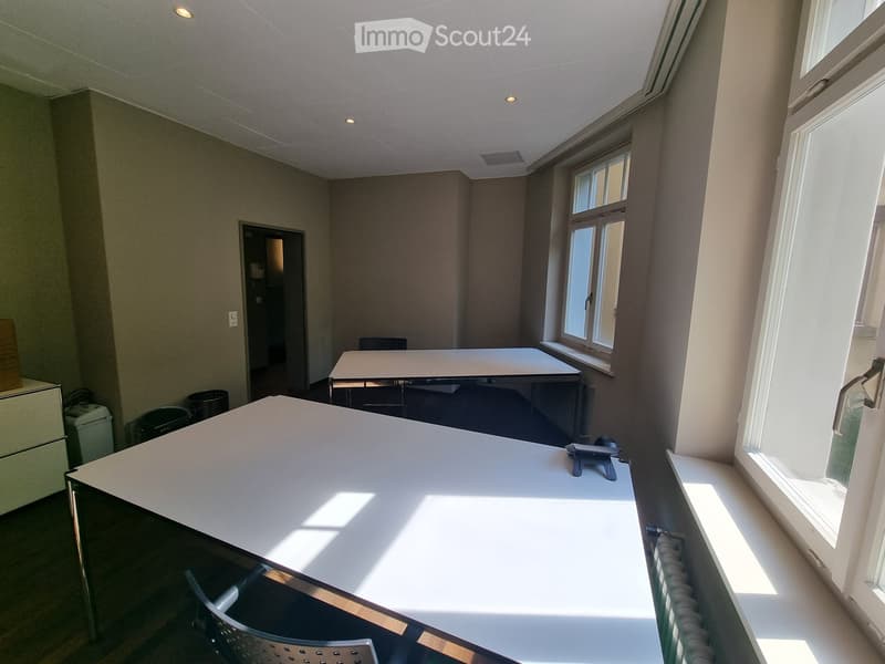 Bright, unfurnished room with large windows, a white table, a chair, and a screen on the table.