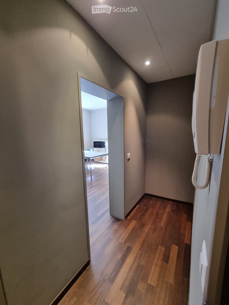 A narrow hallway with wooden flooring and light-colored walls, featuring a partially open door leading to another room with a table and chairs visible in the background.