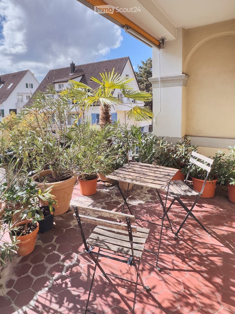 Balcony with wooden table and chairs, surrounded by potted plants and greenery, overlooking a residential area.