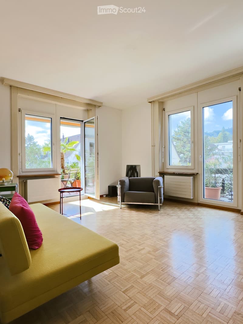 Bright living room with wooden herringbone flooring, large windows, and a view of greenery. Features a green sofa with red pillows, a grey armchair, a small round table, and a decorative ball on a side table.