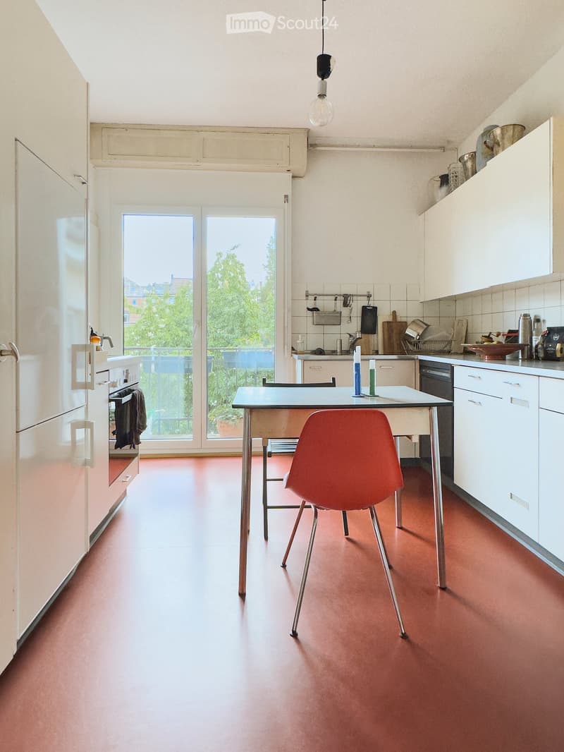 Modern kitchen with white cabinets, red floor, large window, and a small dining area with a red chair and table.