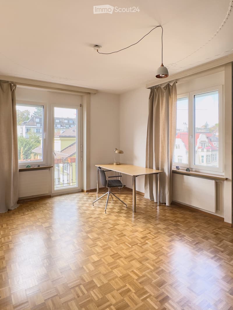 Bright, unfurnished room with wooden herringbone flooring, large windows, and a single desk setup with a chair and lamp.