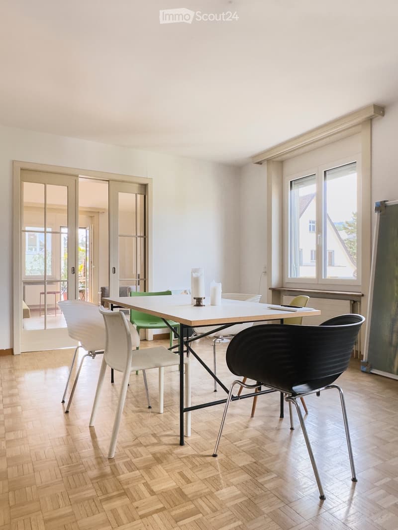 Bright room with wooden herringbone flooring, white table with four chairs, and a candle centerpiece. Large windows and sliding glass doors lead to a balcony area.
