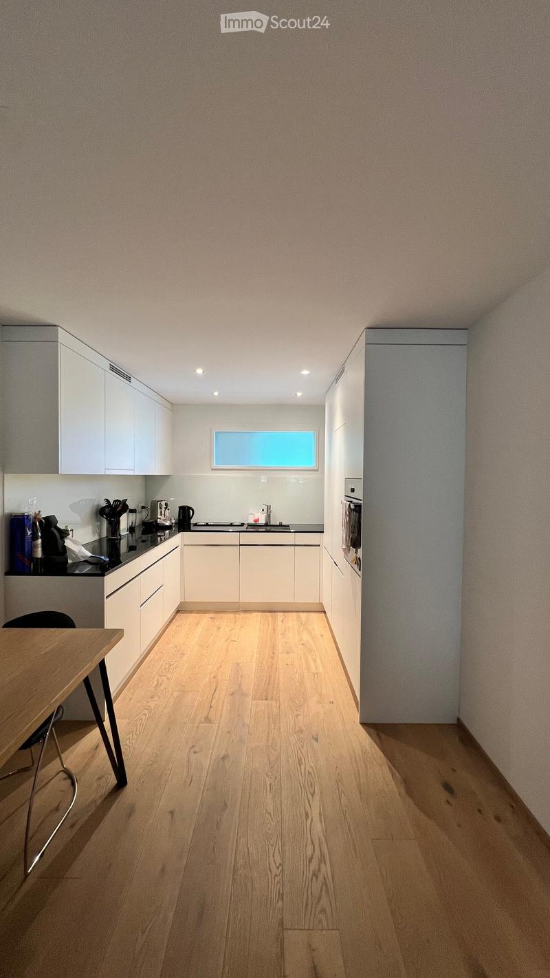 Modern kitchen with light wood flooring, white cabinets, and a black countertop. A small table and chair are positioned near the cabinets, and various kitchen appliances and utensils are visible on the countertop.