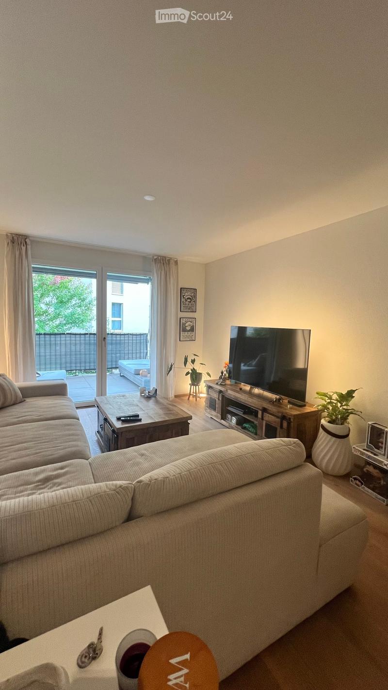 Modern living room with a large sectional sofa, coffee table, flat-screen TV on a wooden stand, and a small plant in a pot. The room features large windows with curtains and a sliding glass door leading to a patio.