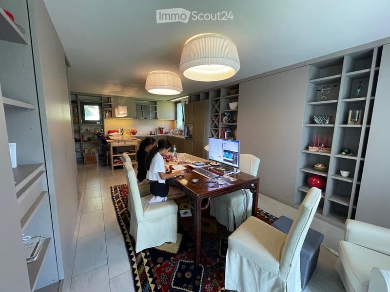 A well-lit room with a large wooden table, chairs, a laptop, and shelves. The room features modern lighting and a cozy rug on the floor.