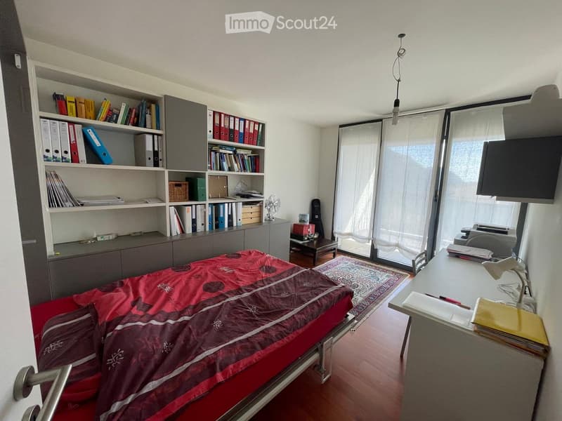 A furnished room with a bed, desk, bookshelf, small table, and various items including a fan, guitar, and printer.