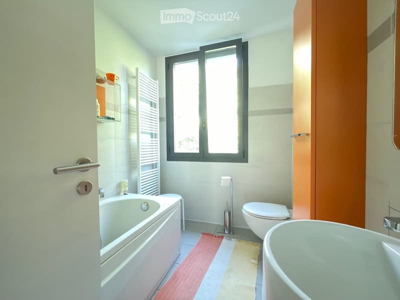 Modern bathroom with white tiles, a bathtub, toilet, towel rack, and a window letting in natural light.