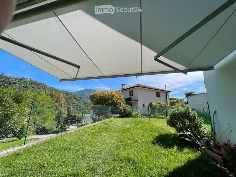Lush green lawn under a large retractable awning, with a white house and mountainous backdrop visible.