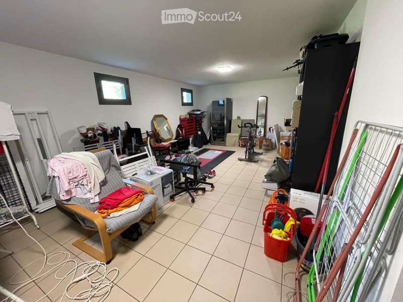 A cluttered room with a tiled floor, a chair with clothes draped over it, a small table with items, a mirror, a fridge, a bicycle, and various storage boxes and cleaning supplies scattered around.