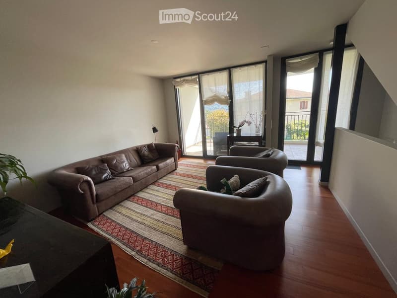 Modern living room with wooden flooring, large windows, and a balcony. Features a brown leather sofa, armchair, coffee table, and decorative plants.