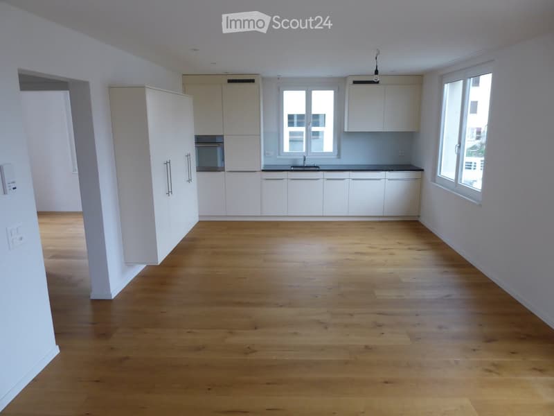 Empty kitchen with white cabinets, wooden flooring, and large windows allowing natural light.