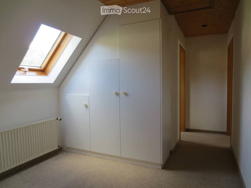 Empty attic room with white built-in wardrobes, skylight window, radiator, and wooden ceiling beams.