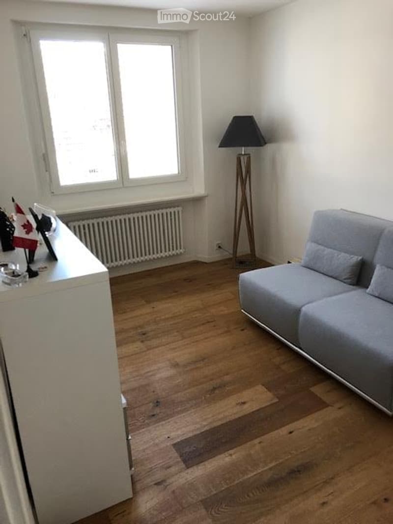 A modern living room with wooden flooring, a grey sofa, a tall floor lamp, and a small table with decorative items near a large window.