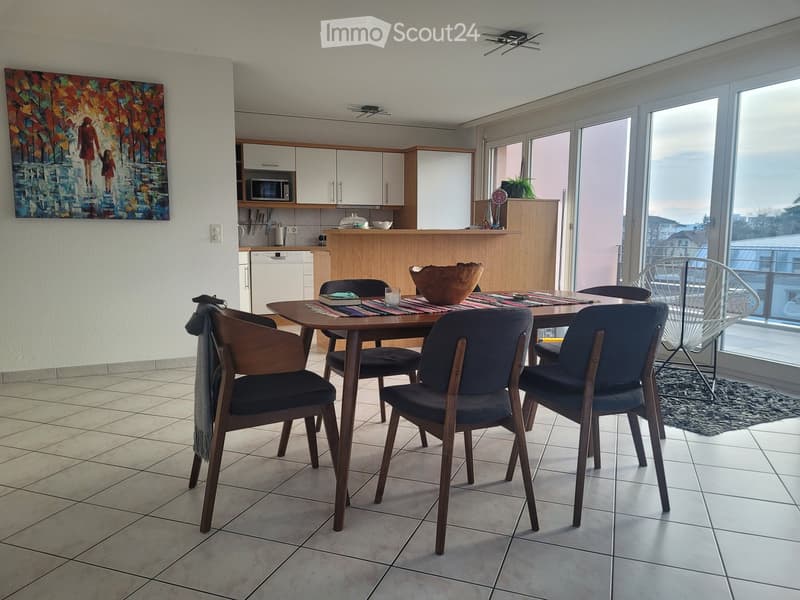 Bright dining area with a wooden table, four chairs, and a modern kitchen in the background. The space features large windows with a view, a colorful painting on the wall, and a small bar area with a microwave and dishwasher.