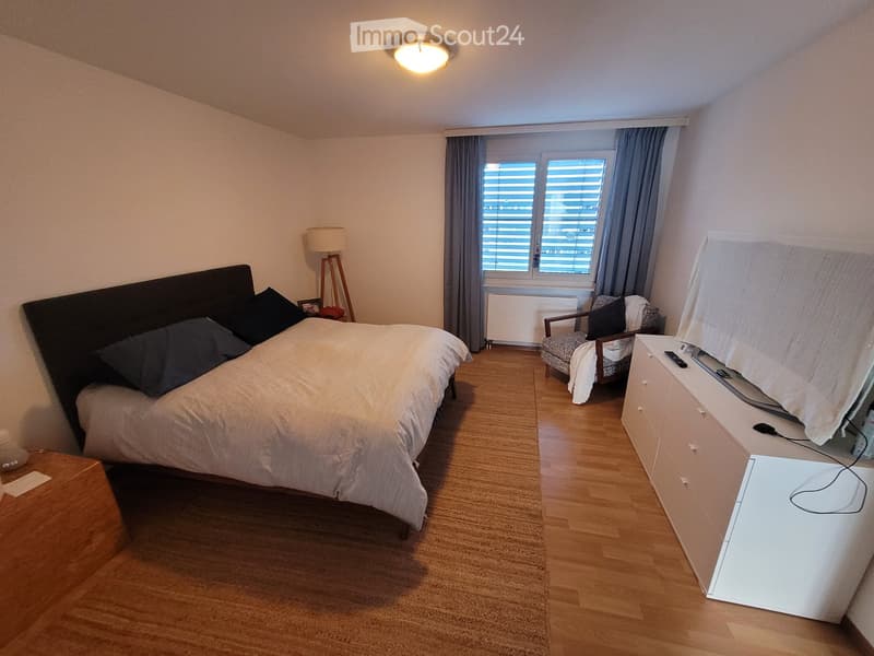 Cozy bedroom with a large bed, nightstand, armchair, dresser, and a lamp. The room features wooden flooring and large windows with blinds.